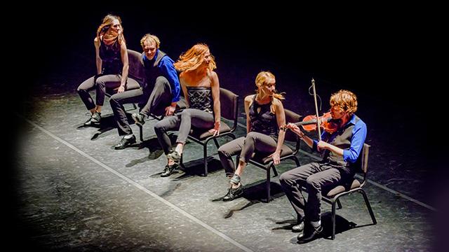 five members of the StepCrew sitting on stage in chairs - one of them is playing a fiddle